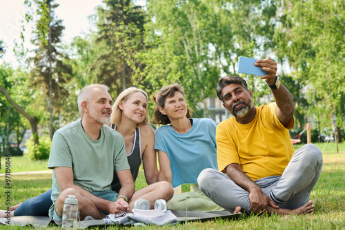 Happy mature people in activewear looking at screen of mobile phone held by multiethnic man while watching online video outdoors