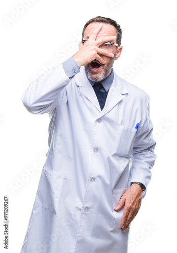 Middle age senior hoary professional man wearing white coat over isolated background peeking in shock covering face and eyes with hand, looking through fingers with embarrassed expression.