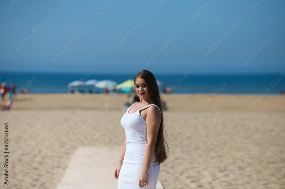 A beautiful young woman with long hair and an elegant white dress walks along the wooden walkway on the sandy beach. In the background the blue sea and the umbrellas of the holidaymakers. A beautiful young woman with long hair and an elegant white dress walks along the wooden walkway on the sandy beach. In the background the blue sea and the umbrellas of the holidaymakers.