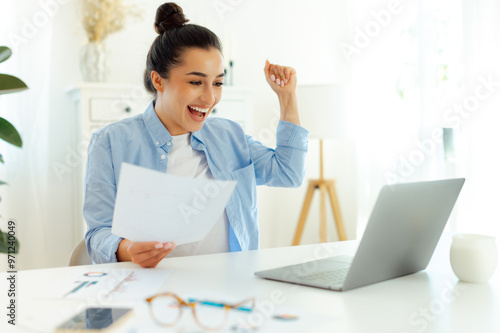 Good job. Cheerful happy indian or arabian successful business woman, sitting at a workplace, joyfully looking at a laptop screen, rejoicing in success, holding documents, smiling, gesturing with fist