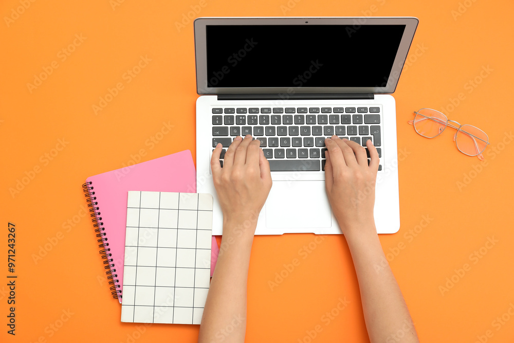 Fototapeta premium Female hands with laptop, notebooks and eyeglasses on orange background