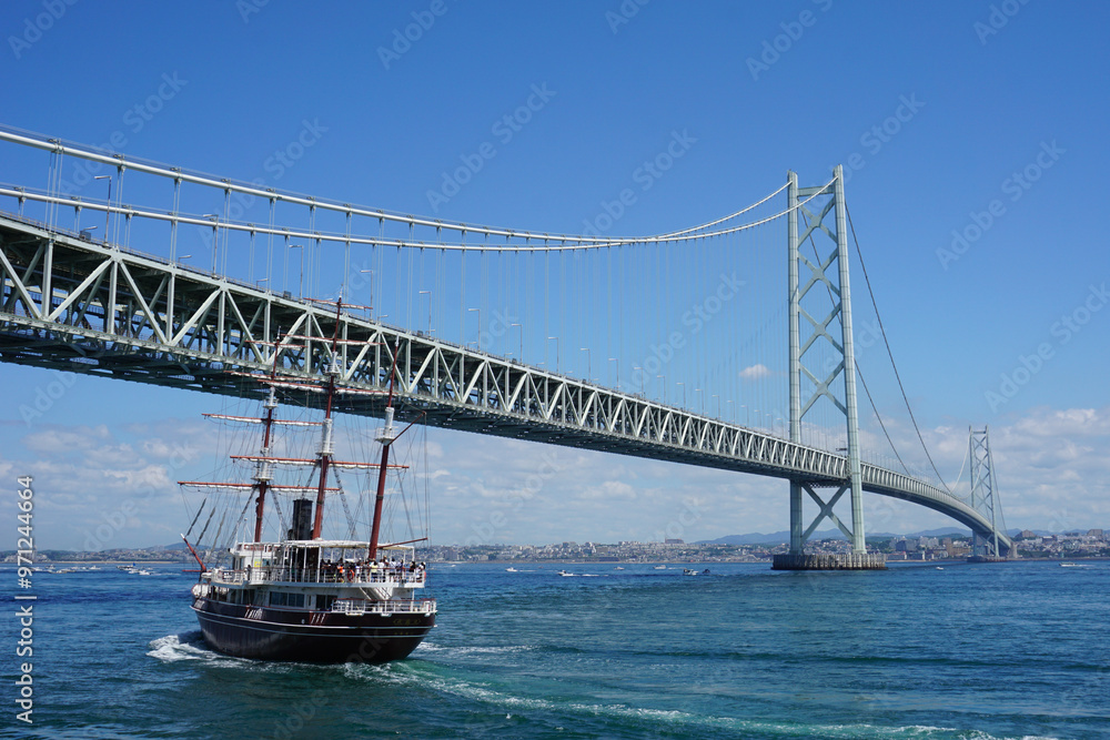 Fototapeta premium 晴天の海にかかる巨大な吊橋と帆船