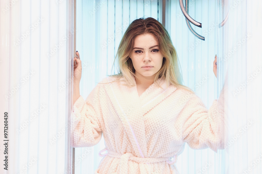 Woman in bathrobe standing in tanning booth with bright lights and calm expression. Sun lifestyle danger. Ultraviolet skin risk. Salon procedure. Female tan equipment Woman in bathrobe standing in tanning booth with bright lights and calm expression. Sun lifestyle danger. Ultraviolet skin risk. Salon procedure. Female tan equipment
