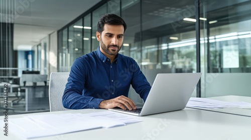 Indian man sitting at an office desk typing on a laptop, focused on work tasks, hybrid work setup, balancing remote and office responsibilities