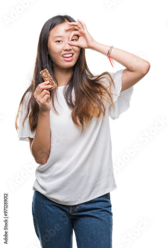 Young asian woman eating chocolate energetic bar over isolated background with happy face smiling doing ok sign with hand on eye looking through fingers