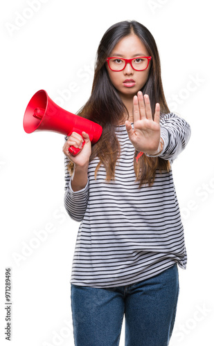 Young asian woman holding megaphone over isolated background with open hand doing stop sign with serious and confident expression, defense gesture