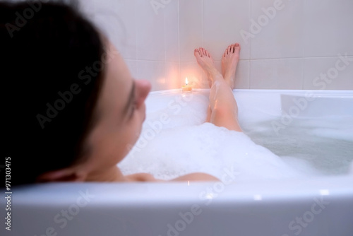 Tired pensive young brunette woman relaxing in bathroom with foam looking at burning candle at home after working day. Resting female enjoying bathing with comfort in apartment.