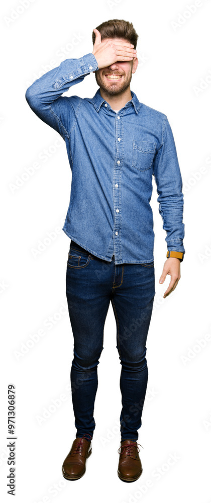 Young handsome blond man wearing casual denim shirt smiling and laughing with hand on face covering eyes for surprise. Blind concept. Young handsome blond man wearing casual denim shirt smiling and laughing with hand on face covering eyes for surprise. Blind concept.