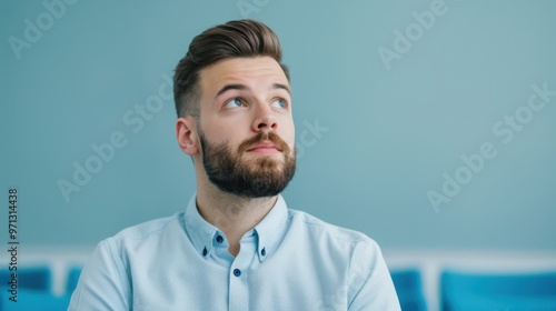 Close-up portrait of a thoughtful young man with a beard, looking up while sitting in a modern office with a soft blue background.