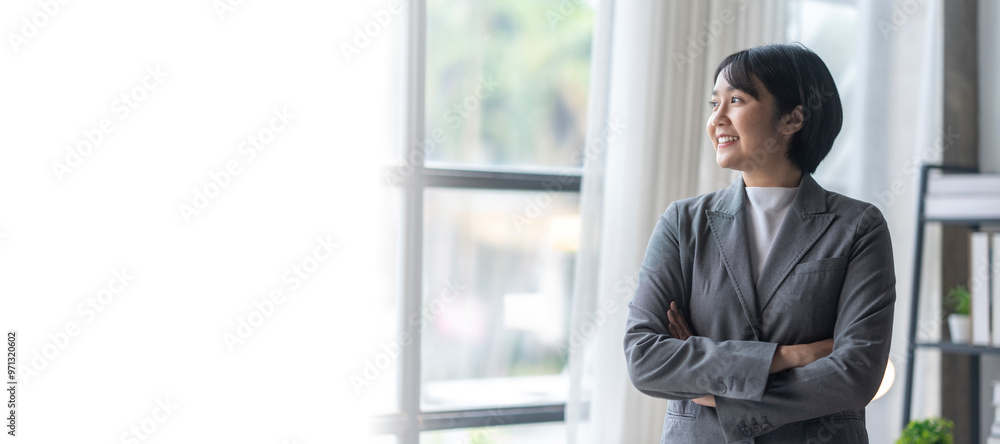 Fototapeta premium Young businesswoman smiling and looking out the window with arms crossed in her office