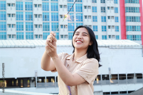Free Download beautiful asian young woman holding sparklers firework and laughing in new year eve celebration standing in roof top outdoor with urban building background - GFXLoad
