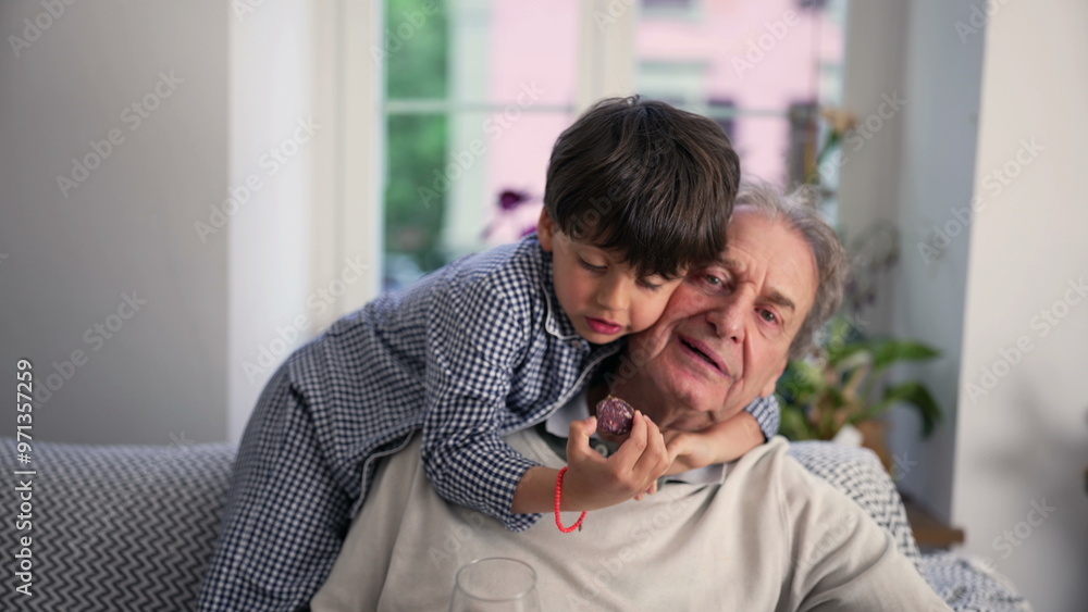 Young boy hugging his grandfather while eating a snack, sharing a moment of affection and bonding on a sofa at home Young boy hugging his grandfather while eating a snack, sharing a moment of affection and bonding on a sofa at home