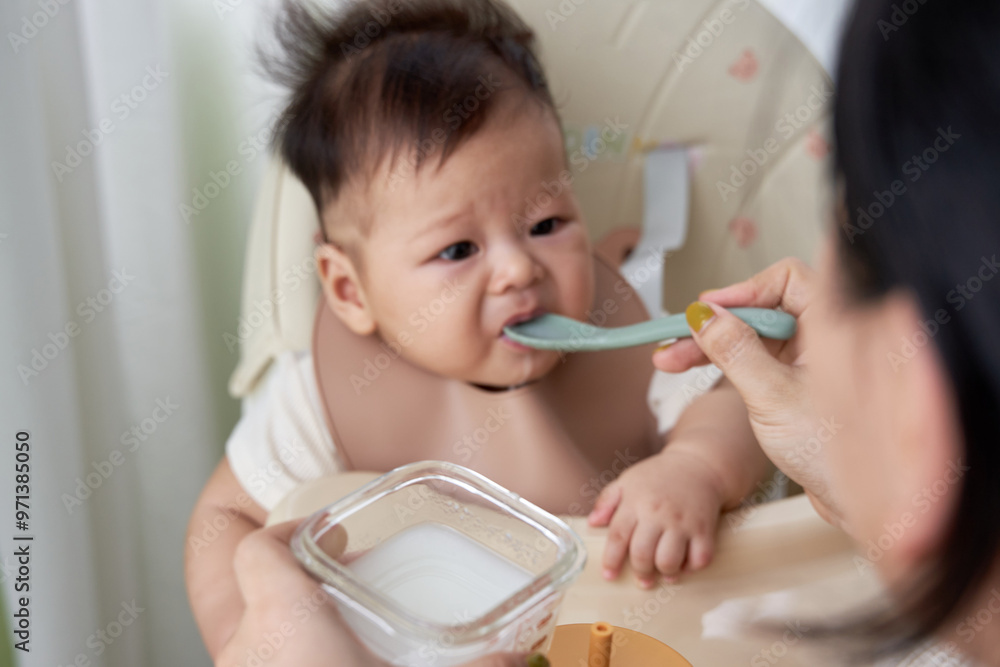 The baby is sitting in a high chair with an indifferent face, when the mother gives the baby food, the light is soft The baby is sitting in a high chair with an indifferent face, when the mother gives the baby food, the light is soft