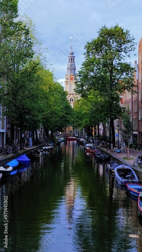 peaceful Amsterdam canal with traditional Dutch houses, parked bicycles, and boats on calm water. Arched bridges and green trees frame the scene, warm sunset light reflecting on historic buildings