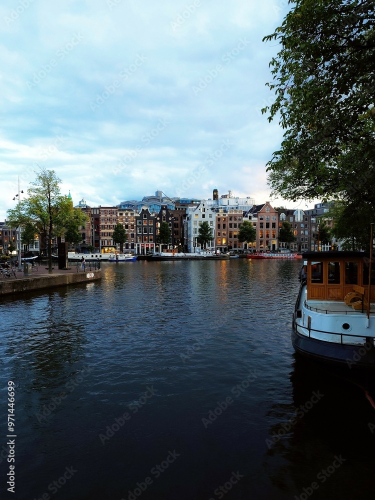 peaceful Amsterdam canal with traditional Dutch houses, parked bicycles, and boats on calm water. Arched bridges and green trees frame the scene, warm sunset light reflecting on historic buildings