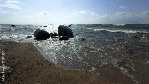 A serene coastal scene featuring a sandy beach with scattered rocks and pebbles along the shoreline, where gentle waves meet the shore under bright sunlight. The water shimmers as it moves across the photo