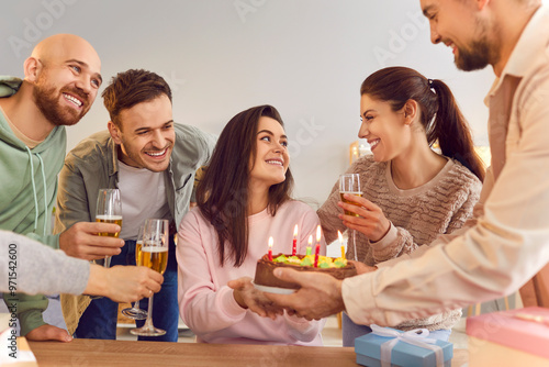 Smiling young people holding glasses of champagne congratulating their happy girl friend sitting at table on her birthday and giving her cake at home. Birthday party and celebration concept.