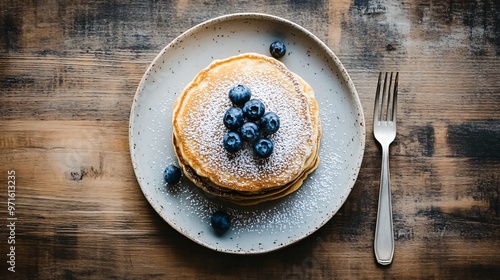 Simple stack of pancakes with blueberries and powdered sugar, placed on a minimalist wooden table with a single piece of cutlery