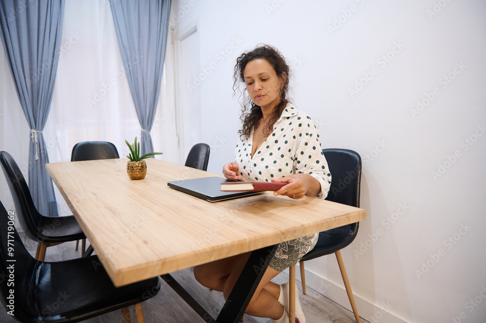 Woman in polka dot blouse sitting at a wooden table with a laptop and notebook in a modern office setting Woman in polka dot blouse sitting at a wooden table with a laptop and notebook in a modern office setting