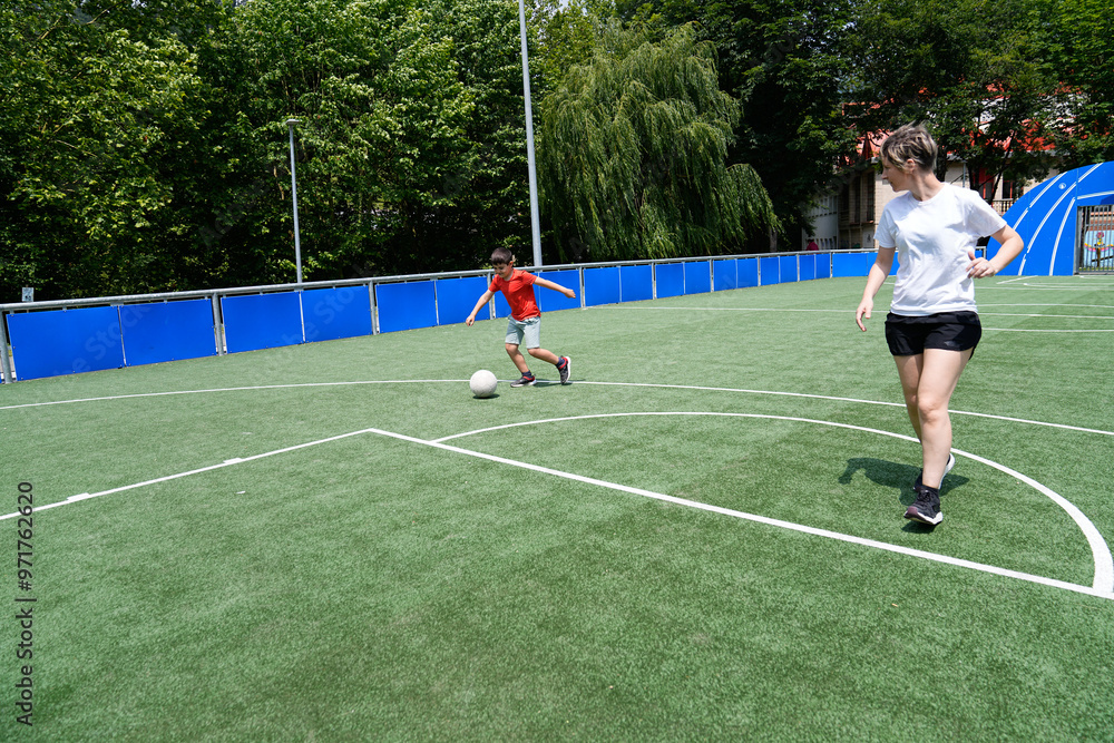 Fototapeta premium Mother and son playing soccer on a bright green field, kicking the ball to each other.