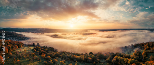Aerial panorama of a misty rural landscape at sunrise, with colorful autumn trees and green meadows, colorful dramatic sky and layers of sunlit mist