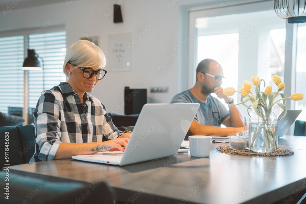 Man and woman sitting by the dining room desk, doing work from home on their laptops Man and woman sitting by the dining room desk, doing work from home on their laptops