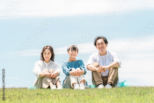 青空の見える公園・丘に座る家族・親子（父親・母親・娘） 