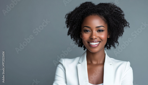 A confident African American businesswoman in a white suit smiles warmly with arms crossed, standing against a neutral background, perfect for corporate or business settings.