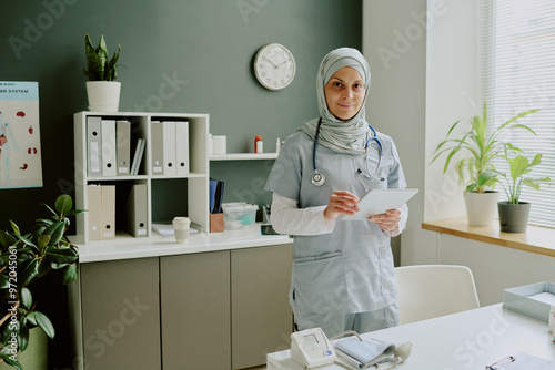 Caucasian woman in medical uniform holding tablet while standing in modern healthcare workspace with various medical devices and plants around photo