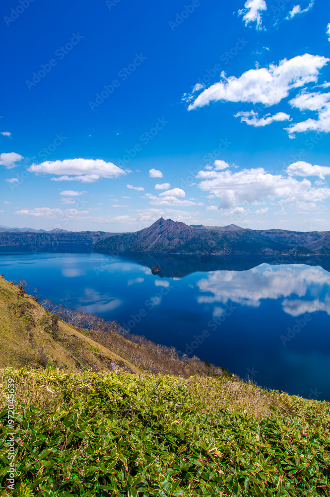 雲の浮かぶ青空を反射する山間の湖。日本の北海道の摩周湖は美しい自然の景色が観光お客に人気の名所。雄大で迫力のあるカルデラ湖の風景が魅力。 雲の浮かぶ青空を反射する山間の湖。日本の北海道の摩周湖は美しい自然の景色が観光お客に人気の名所。雄大で迫力のあるカルデラ湖の風景が魅力。