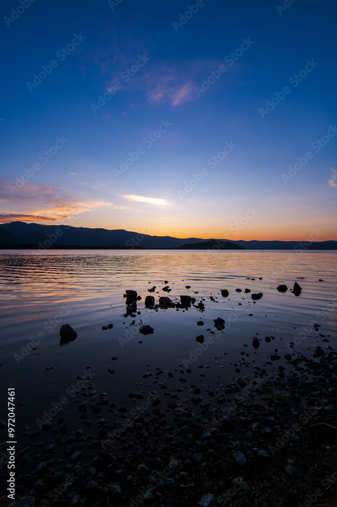 Fototapeta premium 鮮やかなグラデーションの空の色を湖面に反射する夕暮れの湖。岸辺からのローアングル。北海道の屈斜路湖。