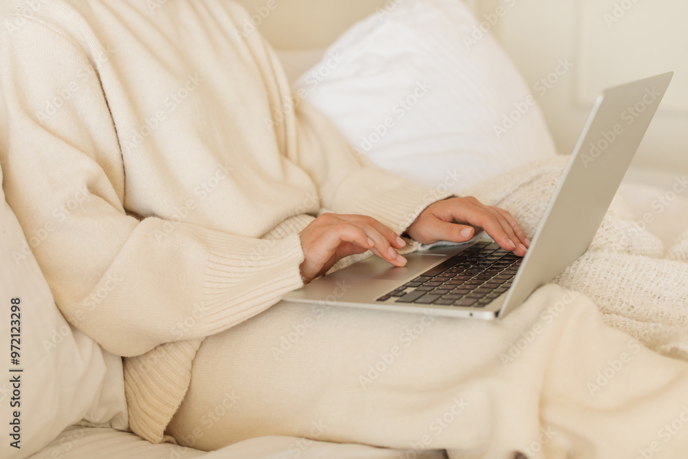 Fototapeta premium Close-up of a young woman in a cozy, light-colored house outfit working on a laptop at home in bed.