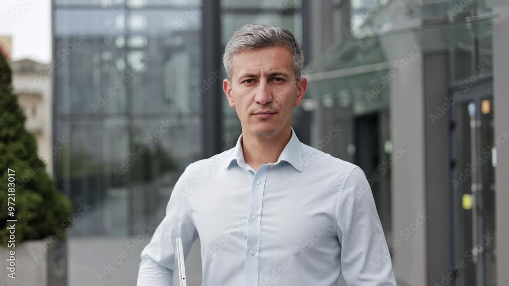 A middle-aged businessman with grey hair, wearing a white shirt, stands outside a modern office building with a laptop in his hand. He has a serious expression on his face, corporate environment. A middle-aged businessman with grey hair, wearing a white shirt, stands outside a modern office building with a laptop in his hand. He has a serious expression on his face, corporate environment.
