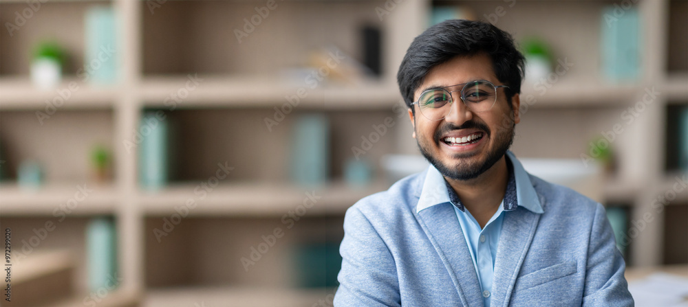 A young Indian man with glasses smiles confidently while sitting in a well-lit modern office. Background features shelves adorned with plants and decorative items, creating a welcoming atmosphere. A young Indian man with glasses smiles confidently while sitting in a well-lit modern office. Background features shelves adorned with plants and decorative items, creating a welcoming atmosphere.