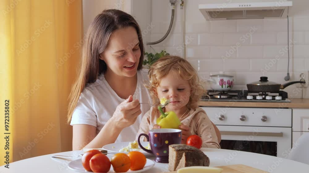 Cheerful mom providing nutritious meal to her cute child enjoying playful moment together at home while having breakfast telling about benefits of vegetables in children's diet Cheerful mom providing nutritious meal to her cute child enjoying playful moment together at home while having breakfast telling about benefits of vegetables in children's diet