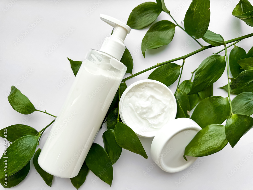 Close up of white lotion bottle and hand cream container with green leaves on white background. Skincare and natural beauty concept. For healthcare, wellness, and organic product poster. Close up of white lotion bottle and hand cream container with green leaves on white background. Skincare and natural beauty concept. For healthcare, wellness, and organic product poster.