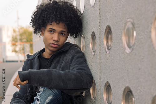 serious african american hipster male teenager with curly hair looking at camera while sitting near concrete wall on urban street, concept of urban lifestyle