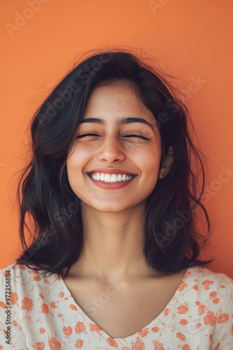 A portrait photo of a cheerful 27-year-old Indian woman winking playfully against a pastel orange background