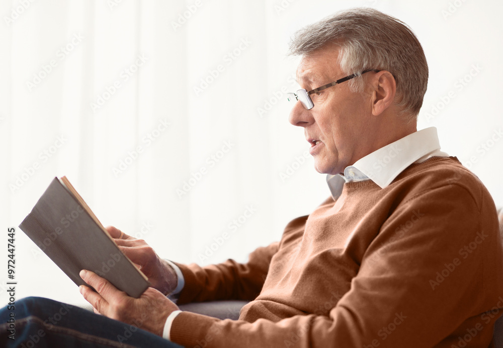 Senior Man Reading Book Sitting On Couch Next To Window At Home On Weekend. Retirement Lifestyle. Selective Focus Senior Man Reading Book Sitting On Couch Next To Window At Home On Weekend. Retirement Lifestyle. Selective Focus
