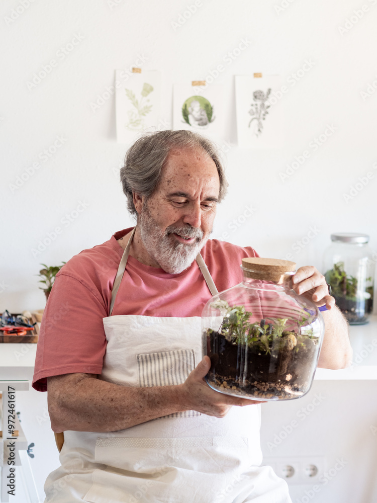 An elderly man with gray hair smiles as he carefully examines the small plants inside a glass terrarium he has made. Sunlight streams through the window of his studio, decorated with more terrariums a An elderly man with gray hair smiles as he carefully examines the small plants inside a glass terrarium he has made. Sunlight streams through the window of his studio, decorated with more terrariums a
