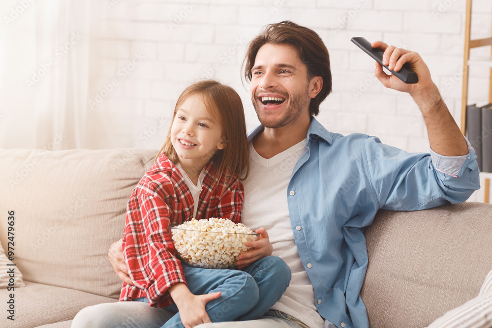 Little girl and her happy father watching football match on TV, celebrating success of their team, sitting on sofa with popcorn, family traditions concept, copy space Little girl and her happy father watching football match on TV, celebrating success of their team, sitting on sofa with popcorn, family traditions concept, copy space