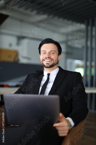 A man in a suit and tie is sitting in a chair with a laptop in front of him. He is smiling and he is happy