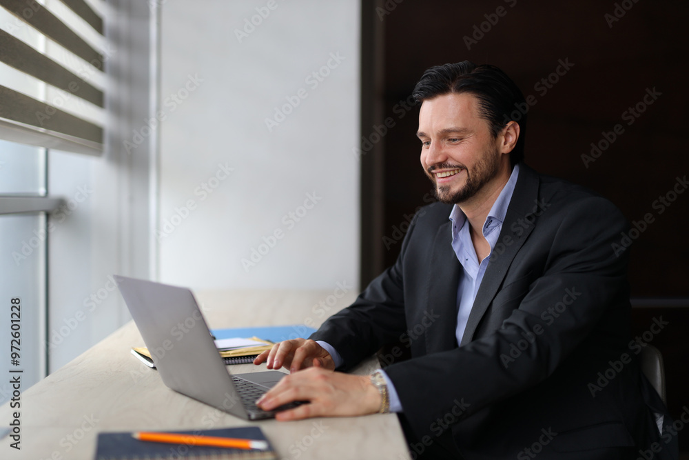 A man in a suit is sitting at a desk with a laptop open in front of him. He is smiling and he is enjoying his work A man in a suit is sitting at a desk with a laptop open in front of him. He is smiling and he is enjoying his work