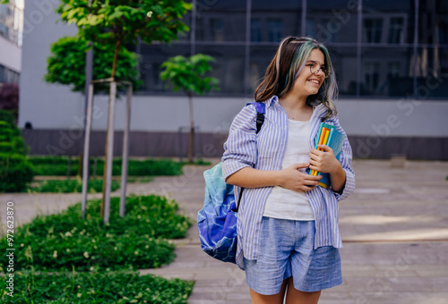 A teenage girl smiling looks away, walking to school dressed in a backpack, carrying notebooks photo