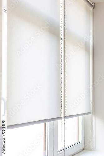 Roller blinds are partially drawn over a window, filtering natural light in a contemporary room, creating a calm and serene ambiance during the day. White roller shades close-up. 