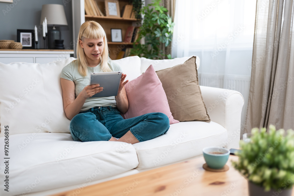 Happy teen girl holding pad computer gadget using digital tablet technology sitting on couch at home. Smiling young woman using apps, shopping, online dating, reading news, browsing internet on sofa. Happy teen girl holding pad computer gadget using digital tablet technology sitting on couch at home. Smiling young woman using apps, shopping, online dating, reading news, browsing internet on sofa.