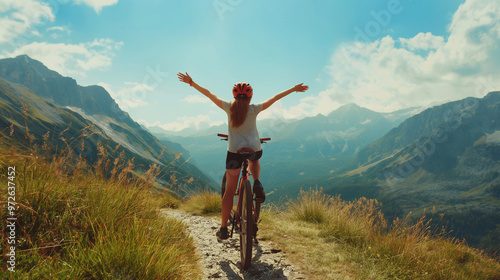 Woman on bicycle raises her arms joyfully, embracing the breathtaking mountain landscape under a bright sky during the day. The scene captures a sense of freedom and adventure