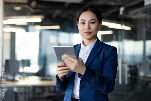 Confident Asian businesswoman using digital tablet in contemporary office setting. Demonstrates concept of technology, communication, and professional productivity in corporate environment