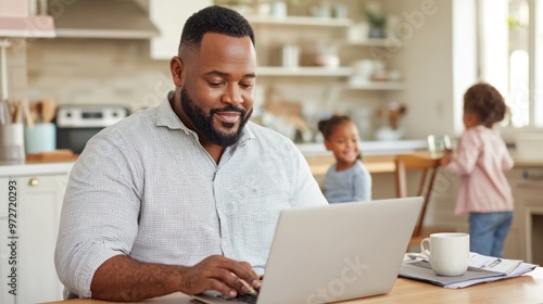 Smiling plus-size black father working on laptop at home with children in the kitchen