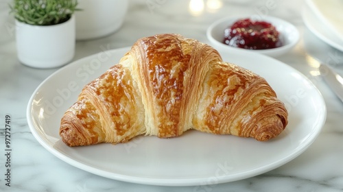 A perfectly shaped croissant on a ceramic plate, served with a side of jam and butter, on a marble countertop.